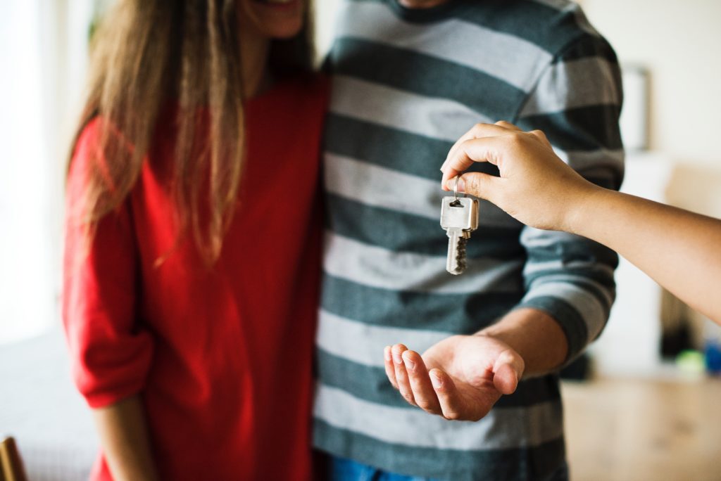 Young couple standing in new house; young male reaching out hand as realtor hands him keys.
