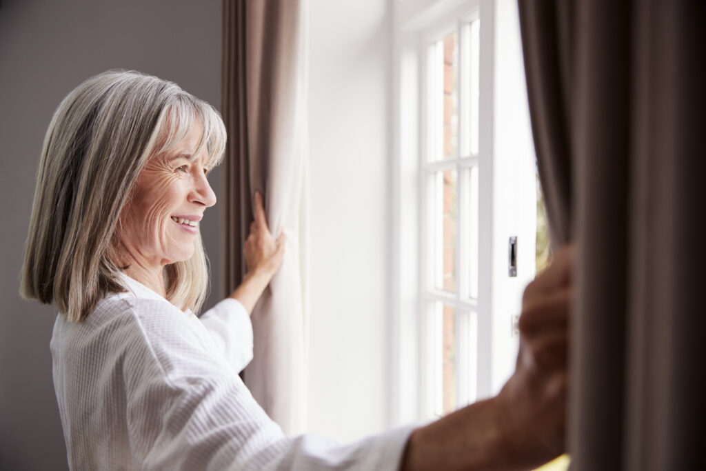 A smiling woman opening the curtains and looking out of the window