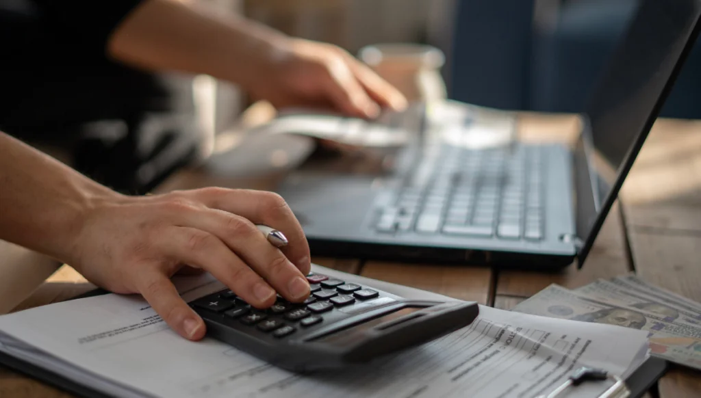 Close-up of someone's hand on a calculator, on top of a stack of papers on a desk, with laptop in background with other hand on keyboard.